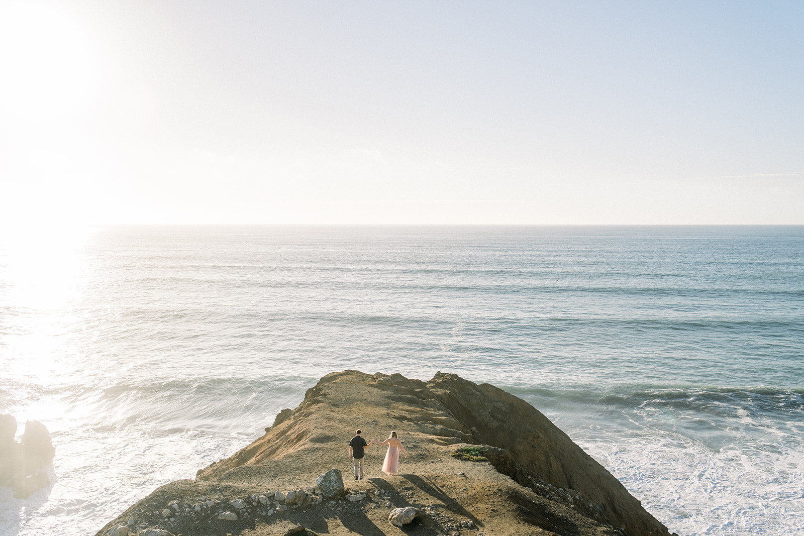 Engaged couple standing on a rocky coastal bluff above the Pacific Ocean at golden hour on the Bay Area coast.