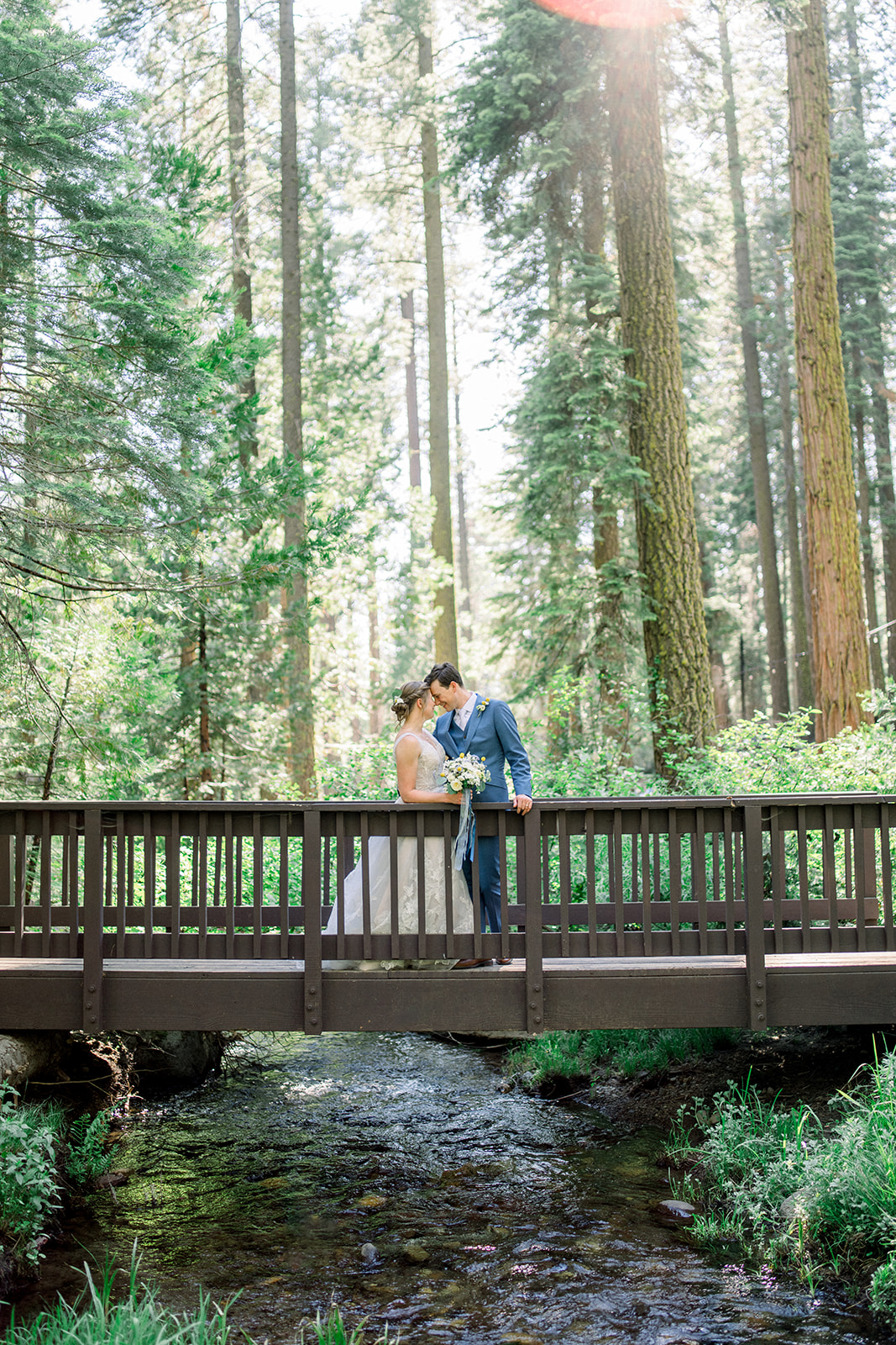 Bride and groom embracing on a wooden footbridge over a creek surrounded by tall pines at Pinecrest Chalet