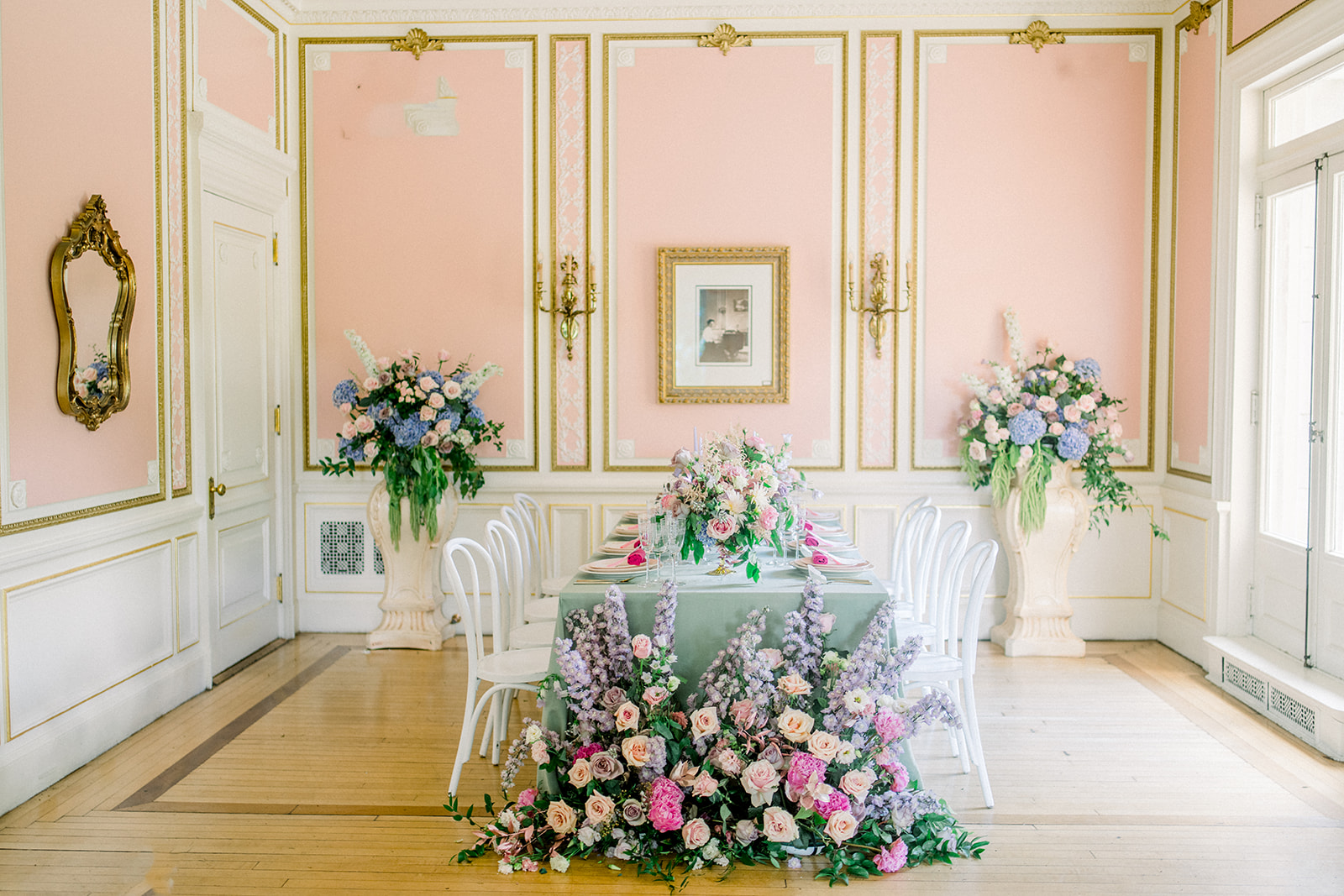 Exquisite wedding reception setup at Cairnwood Estate with a lavish floral centerpiece, elegant white chairs, and ornate gold mirrors accenting the pastel pink walls.