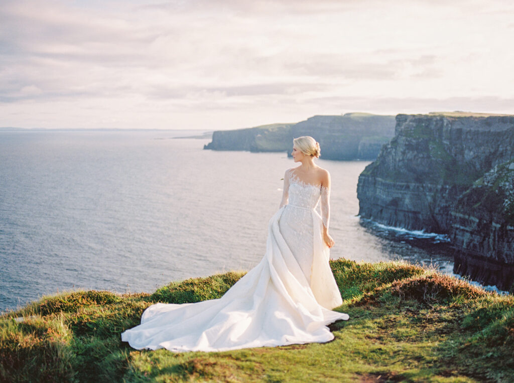 Bride in an elegant lace gown standing on the Cliffs of Moher, Ireland, overlooking the Atlantic Ocean, showcasing the beauty of a destination wedding in a dramatic coastal setting.