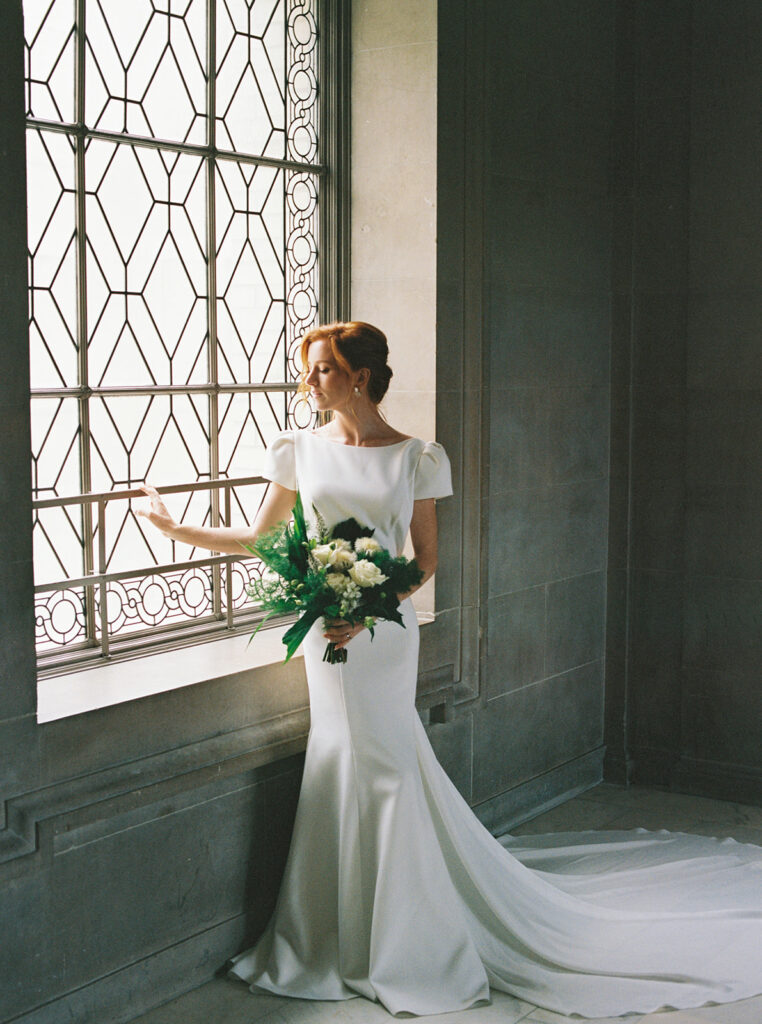 A film capture of a bride standing by a large window at  her San Francisco City Hall Wedding while holding a bouquet of white flowers, in a sleek, minimalist wedding gown, showcasing an elegant and timeless pose in a softly lit, classic interior.