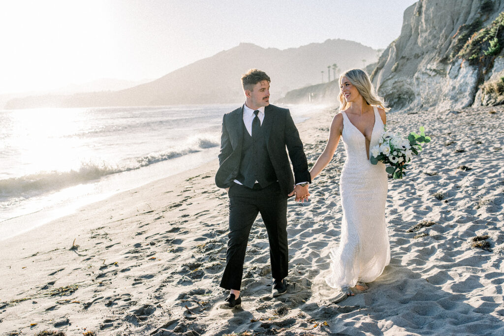 Bride and groom walk hand-in-hand along the beach during their Pismo Beach wedding, with the ocean and cliffs at Dolphin Bay Resort in the background.