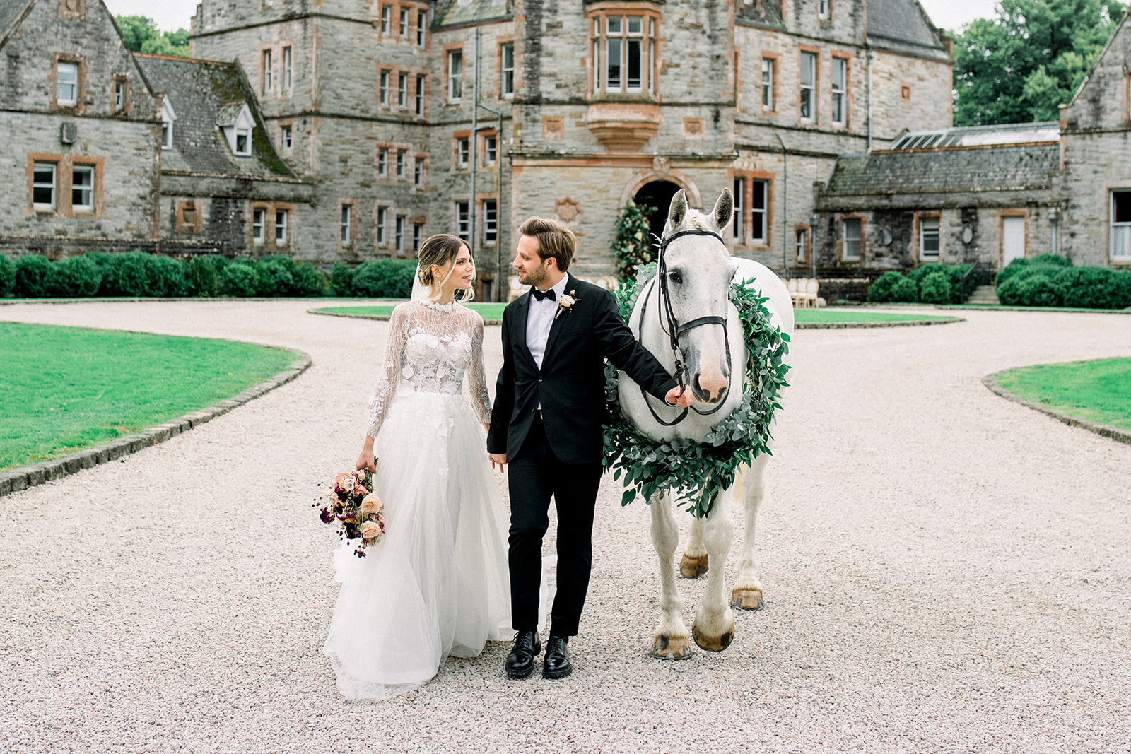 Bride and groom walking with a white horse adorned with greenery at Castle Leslie during their Ireland destination wedding