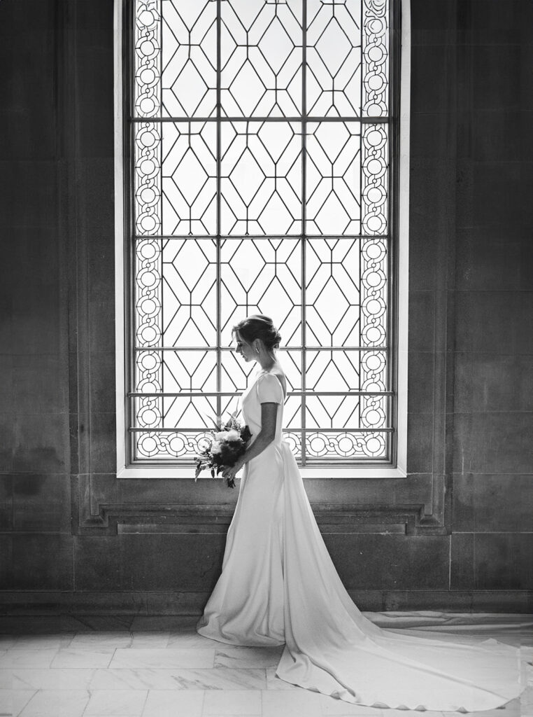 Bride posing by a large, intricately designed window inside San Francisco City Hall, showcasing her wedding gown at her San Francisco wedding.