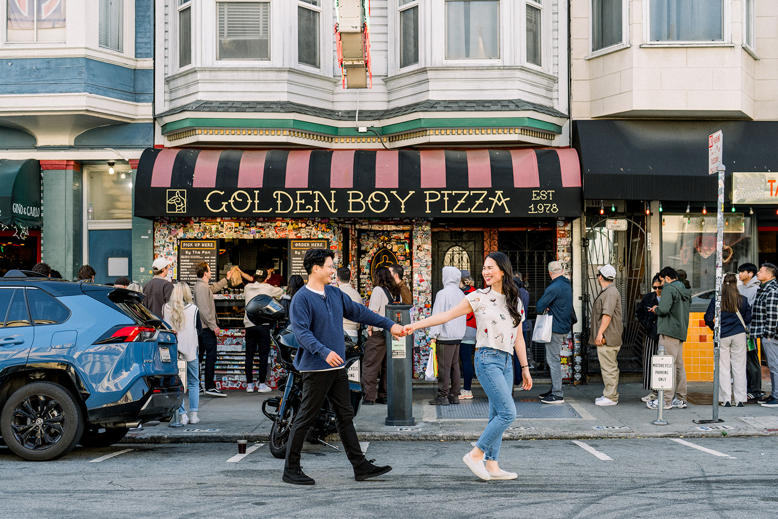 Couple holding hands and crossing the street in front of Golden Boy Pizza during a North Beach engagement session in San Francisco