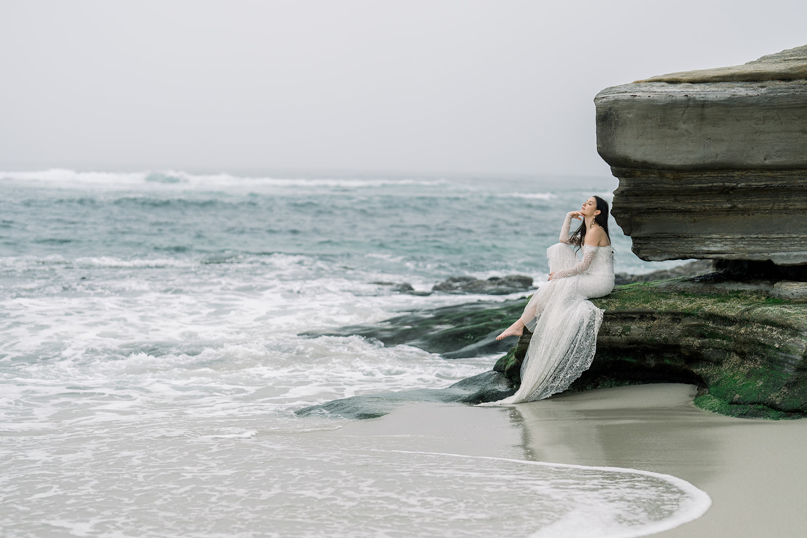 Bride in a lace wedding gown seated on a coastal rock with ocean waves behind her, captured by California wedding photographer