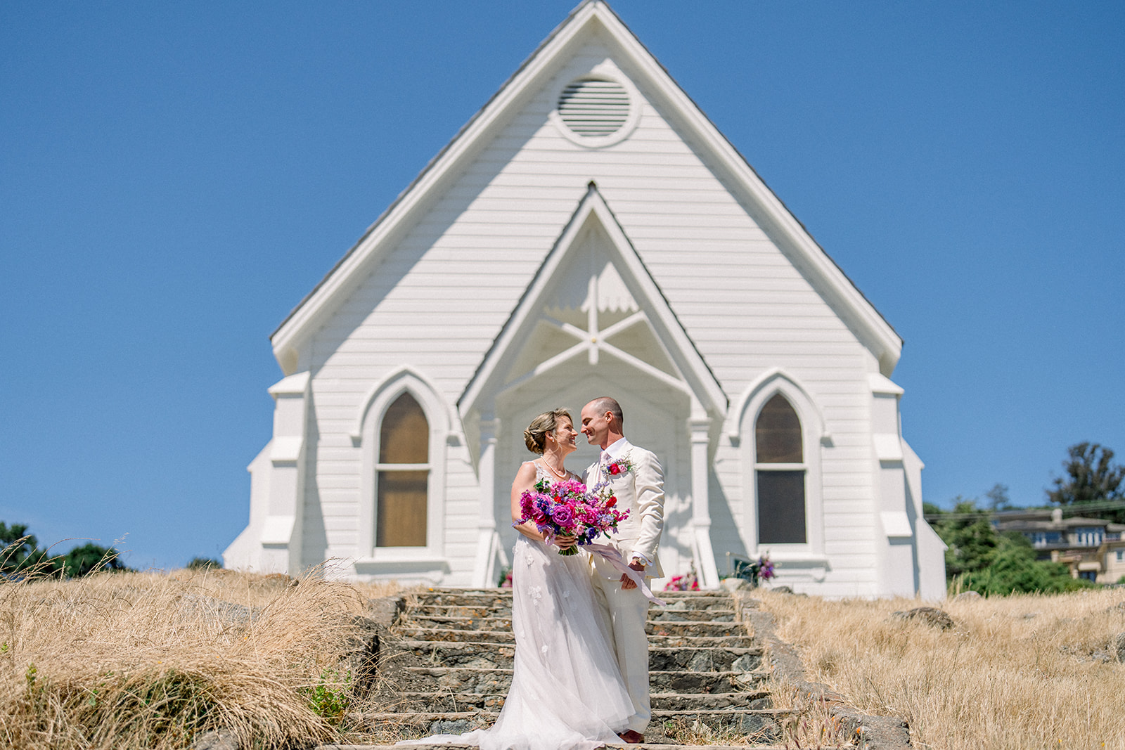 Bride and groom share a joyful moment on the stone steps of Old St. Hilary’s Chapel during their Corinthian Yacht Club wedding in Tiburon, California.