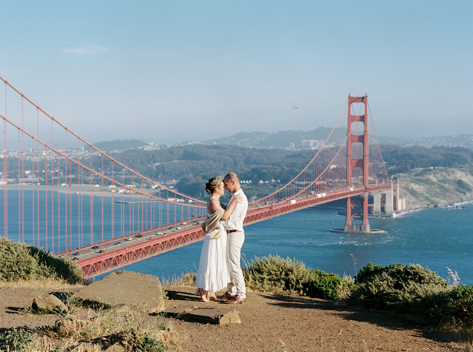 Engaged couple standing at Battery Spencer Outlook during golden hour with the Golden Gate Bridge and San Francisco skyline in the background