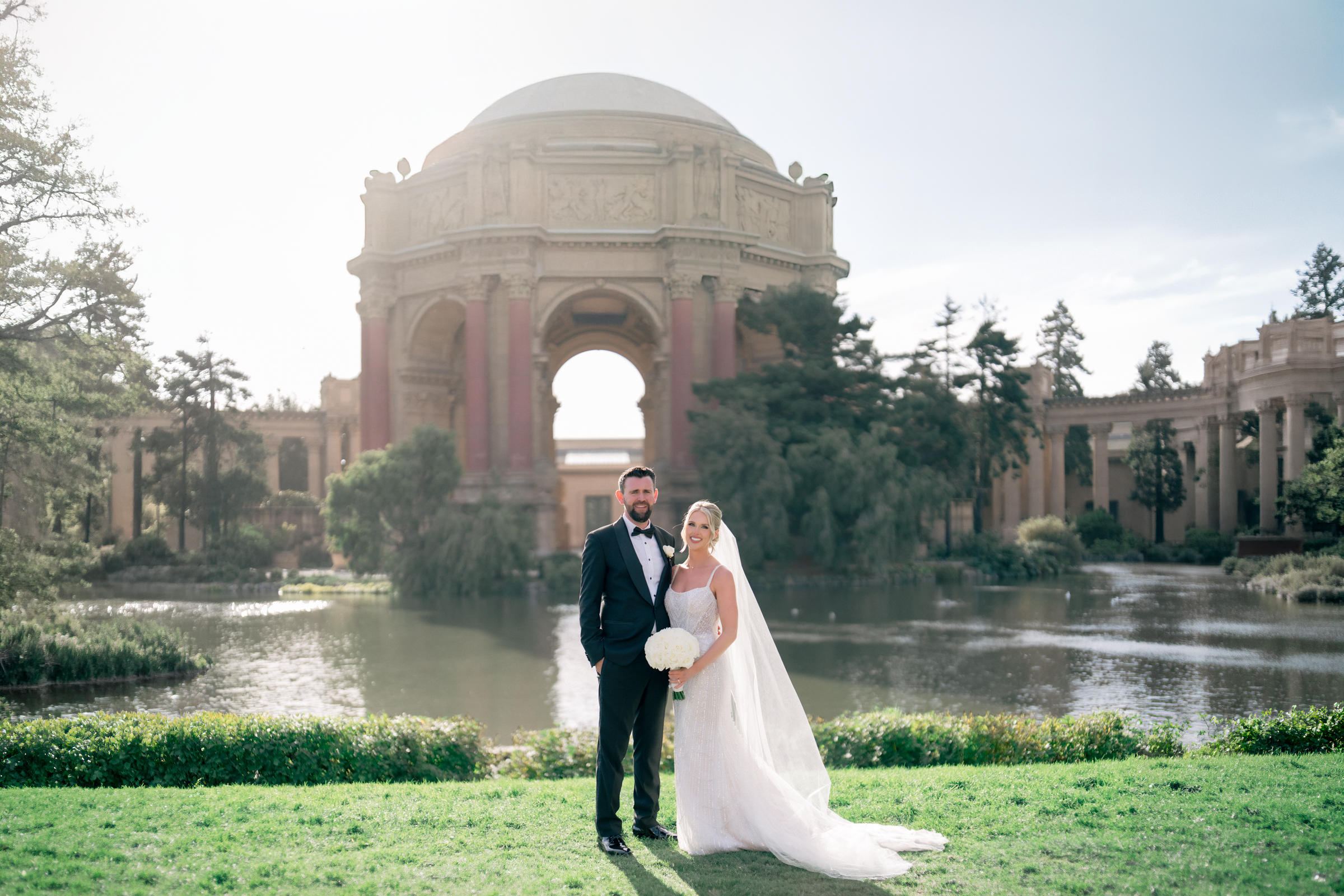 Bride and groom standing on the lawn by the lagoon with the Palace of Fine Arts Rotunda behind them on their San Francisco wedding day